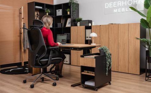Kobieta siedząca w ergonomicznym fotelu przy dębowym biurku z czarnym stelażem. Obok biurka znajduje się czarny kontener na kółkach z dębowym frontem, a w tle widać nowoczesne regały biurowe i rośliny doniczkowe.