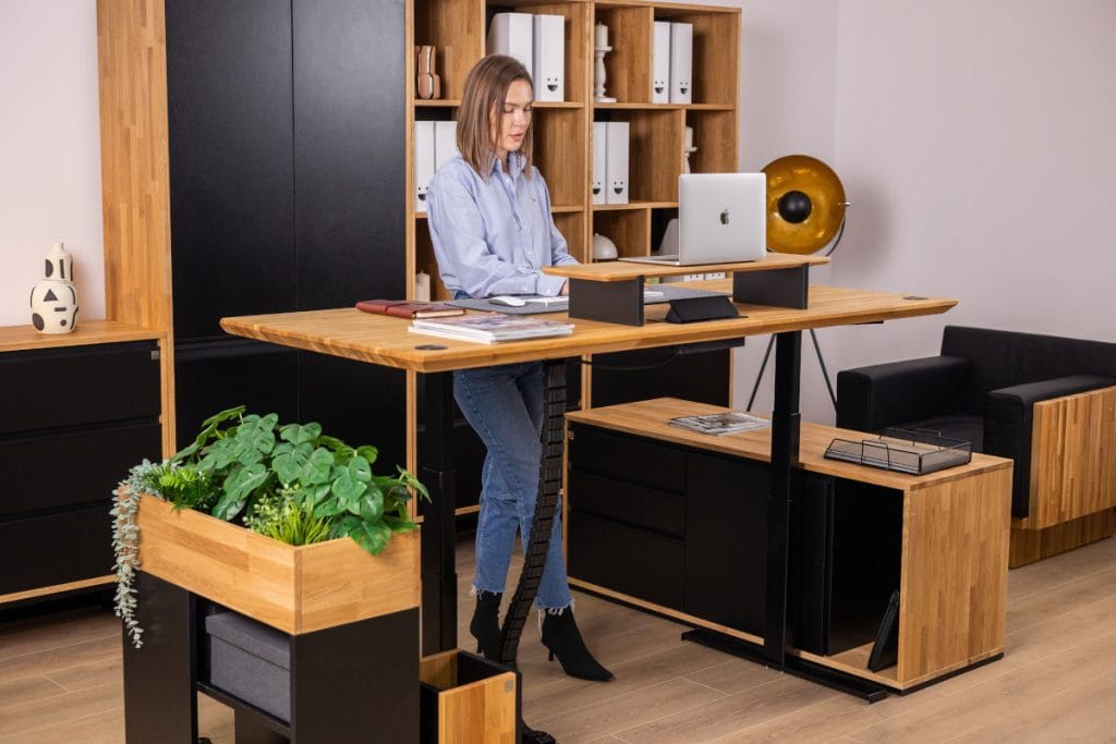 Kobieta korzystająca z biurka do pracy stojącej z dębowym blatem i czarnym stelażem. Na blacie znajduje się nadstawka pod laptopa. Pod biurkiem widoczny pionowy organizer kabli w formie czarnego kręgosłupa.