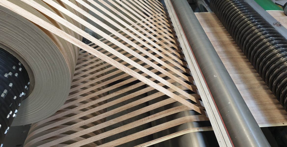 Zbliżenie na przemysłową maszynę do nakładania obrzeży meblowych w trakcie pracy. Widać dużą rolkę jasnego obrzeża PVC z wzorem drewna, które jest precyzyjnie prowadzone przez system rolek i dociskane do krawędzi płyty meblowej. Profesjonalny proces okleinowania wąskich płaszczyzn w fabryce mebli.