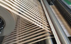 Zbliżenie na przemysłową maszynę do nakładania obrzeży meblowych w trakcie pracy. Widać dużą rolkę jasnego obrzeża PVC z wzorem drewna, które jest precyzyjnie prowadzone przez system rolek i dociskane do krawędzi płyty meblowej. Profesjonalny proces okleinowania wąskich płaszczyzn w fabryce mebli.