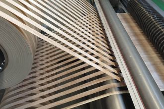 Zbliżenie na przemysłową maszynę do nakładania obrzeży meblowych w trakcie pracy. Widać dużą rolkę jasnego obrzeża PVC z wzorem drewna, które jest precyzyjnie prowadzone przez system rolek i dociskane do krawędzi płyty meblowej. Profesjonalny proces okleinowania wąskich płaszczyzn w fabryce mebli.