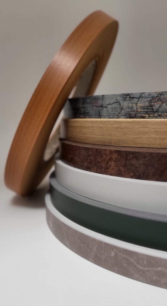 Aranżacja rolek obrzeży meblowych na białym tle, przedstawiająca paletę trendów kolorystycznych. Obok centralnego stosu z różnymi dekorami (drewno, kamień, kolory uni), oparta jest jedna luźna rolka w odcieniu ciepłego drewna, eksponująca bok i strukturę materiału. Prezentacja oferty dla stolarzy i producentów mebli.