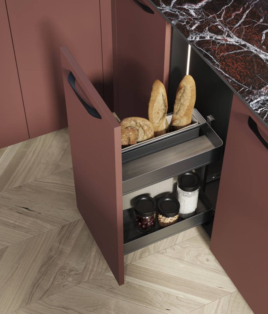 Widok z góry na otwartą szufladę w dolnej szafce kuchennej z systemem wewnętrznych przegródek. Wewnątrz znajdują się dwie świeże bagietki w metalowym koszyku oraz szklane słoiki z kawą i bakaliami.