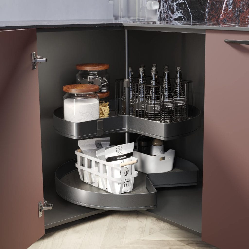 Otwarta szafka narożna typu "nerka" (Le Mans) w dolnej zabudowie kuchennej. Wysunięte półki w ciemnym wykończeniu z wkładem drewnopodobnym, na których znajdują się puszki z napojami, słoiki z mąką, makaronem oraz białym koszykiem z produktami spożywczymi.