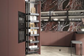Otwarta wysoka szafka cargo w nowoczesnej kuchni z frontami w odcieniu rdzawej czerwieni i blatem z ciemnego marmuru. Wielopoziomowy system wysuwny z koszami wypełnionymi zapasami jedzenia, słoikami, torebkami makaronu i puszkami.