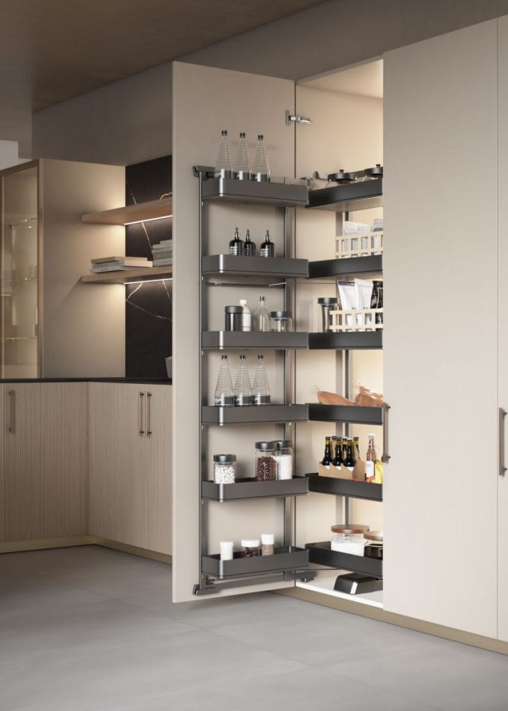 Otwarta wysoka szafka typu cargo w nowoczesnej, jasnej kuchni, stanowiąca system domowej spiżarni. Wielopoziomowe kosze z oświetleniem LED, wypełnione różnorodnymi produktami w butelkach, puszkach i pudełkach.