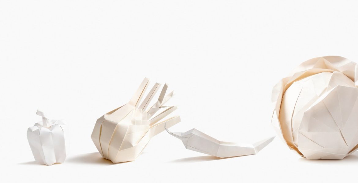 Cztery białe papierowe rzeźby wykonane techniką origami, przedstawiające warzywa ułożone w rzędzie na białym tle. Od lewej: mała papryka, czosnek lub cebula, papryczka chili oraz duża główka kapusty.