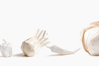 Cztery białe papierowe rzeźby wykonane techniką origami, przedstawiające warzywa ułożone w rzędzie na białym tle. Od lewej: mała papryka, czosnek lub cebula, papryczka chili oraz duża główka kapusty.