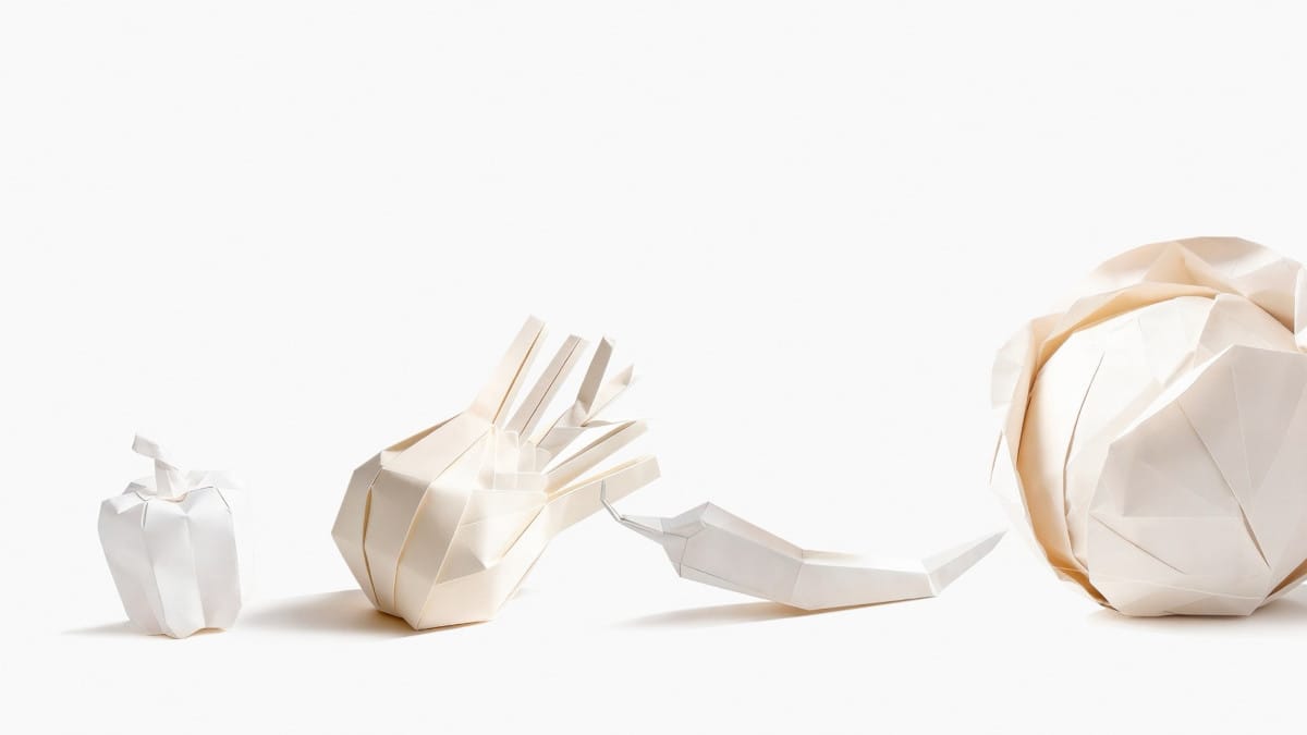 Cztery białe papierowe rzeźby wykonane techniką origami, przedstawiające warzywa ułożone w rzędzie na białym tle. Od lewej: mała papryka, czosnek lub cebula, papryczka chili oraz duża główka kapusty.