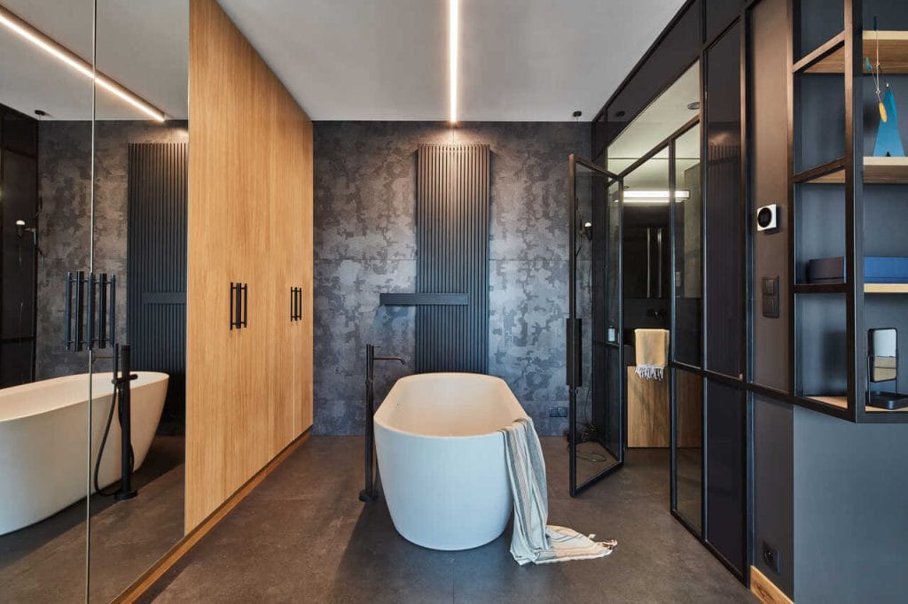 Łazienka na antresoli (druga w tym wnętrzu). Wanna znajduje się w sypialni. Szyby mają przyciemnioną ramę (w formie odwróconej litery „U”). Góra tej ramy skrywa klimatyzację kanałową dla tej przestrzeni, natomiast dzięki pionowym elementom (lewym i prawym) z łóżka widać toalety. Dzięki szklanej przegrodzie łazienka łączy się przestrzennie z sypialnią. Projekt: arch. Roland Stańczyk, RS Studio Projektowe. Fot. Budzik Studio.