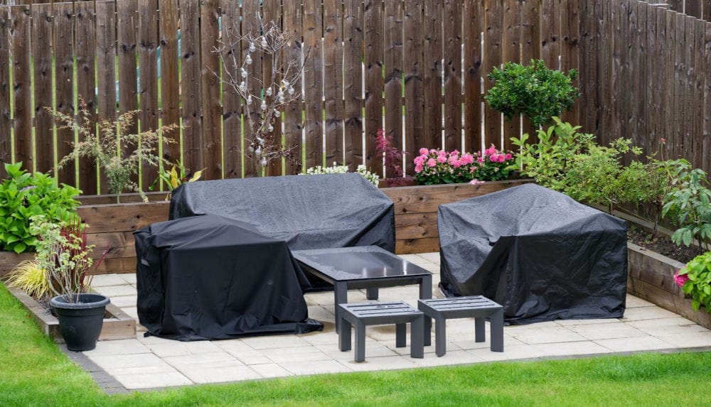 Komplet mebli ogrodowych (kanapa + dwa fotele) zakryte, zabezpieczone ciemną plandeką przed deszczem.