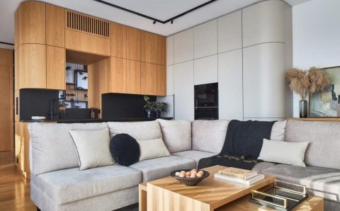 Jednoprzestrzenna kuchnia połączona z salonem. Naturalny dąb połączony z czarnym granitem, który dobrze sprawdza się na blatach. W kuchni jest otwór w zabudowie, łączący kuchnię z klatką schodową. Otwór powstał w wyniku zabudowy obłym elementem meblowym słupa (taka była konstrukcja budynku). Projektant zostawił otwór, aby osoba pracująca w kuchni miała komunikację z przestrzenią klatki schodowej, gdzie się otwiera widok na dwie kondygnacje. Projekt: arch. Roland Stańczyk, RS Studio Projektowe. Fot. Budzik Studio.