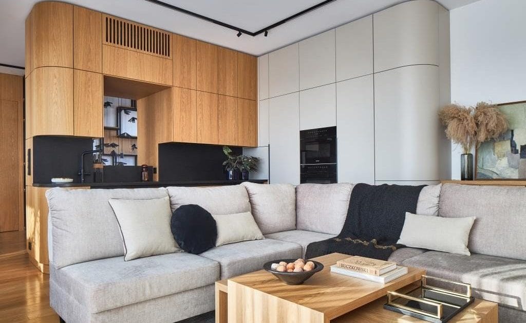 Jednoprzestrzenna kuchnia połączona z salonem. Naturalny dąb połączony z czarnym granitem, który dobrze sprawdza się na blatach. W kuchni jest otwór w zabudowie, łączący kuchnię z klatką schodową. Otwór powstał w wyniku zabudowy obłym elementem meblowym słupa (taka była konstrukcja budynku). Projektant zostawił otwór, aby osoba pracująca w kuchni miała komunikację z przestrzenią klatki schodowej, gdzie się otwiera widok na dwie kondygnacje. Projekt: arch. Roland Stańczyk, RS Studio Projektowe. Fot. Budzik Studio.