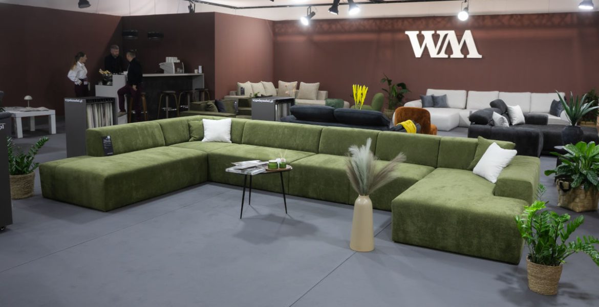 Przestronne stoisko targowe z nowoczesnymi, modułowymi sofami. Na pierwszym planie biała, puszysta sofa narożna w kształcie litery L z żółtymi poduszkami. Z tyłu widać drugą białą sofę oraz szare sofy w tle. Na ścianie logo "wygodnymebel.pl".