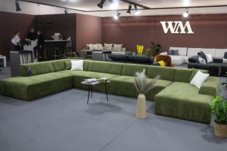 Przestronne stoisko targowe z nowoczesnymi, modułowymi sofami. Na pierwszym planie biała, puszysta sofa narożna w kształcie litery L z żółtymi poduszkami. Z tyłu widać drugą białą sofę oraz szare sofy w tle. Na ścianie logo "wygodnymebel.pl".