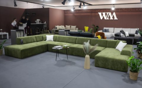 Przestronne stoisko targowe z nowoczesnymi, modułowymi sofami. Na pierwszym planie biała, puszysta sofa narożna w kształcie litery L z żółtymi poduszkami. Z tyłu widać drugą białą sofę oraz szare sofy w tle. Na ścianie logo "wygodnymebel.pl".