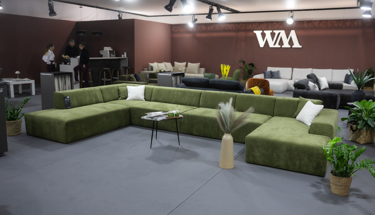 Przestronne stoisko targowe z nowoczesnymi, modułowymi sofami. Na pierwszym planie biała, puszysta sofa narożna w kształcie litery L z żółtymi poduszkami. Z tyłu widać drugą białą sofę oraz szare sofy w tle. Na ścianie logo "wygodnymebel.pl".