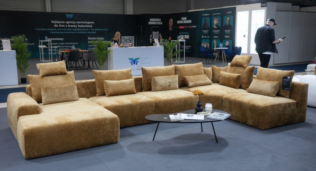 Ogromna, modułowa sofa narożna w kolorze musztardowym lub jasnobrązowym, pokryta miękkim, welurowym materiałem, z licznymi poduszkami. Przed nią niski, okrągły stolik kawowy. W tle widać bannery reklamowe.