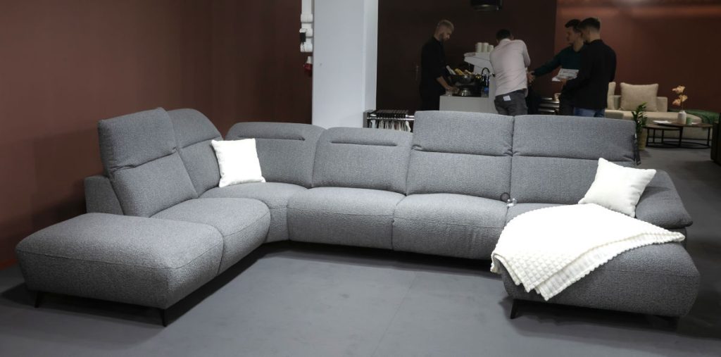 Szeroka, modułowa sofa narożna w kolorze szarym, wykonana z materiału o ciekawej teksturze, z białymi poduszkami. W tle widać pracowników stoiska i klientów stojących przy ladzie barowej.