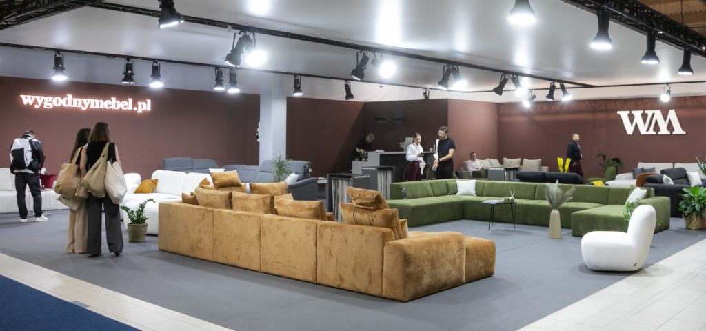 Szeroki kadr stoiska targowego. Na pierwszym planie długa, modułowa sofa w kolorze musztardowego weluru. W tle widoczne są inne sofy (zielone, szare) i strefa barowa, a na ścianie logo "wygodnymebel.pl" i "WM".