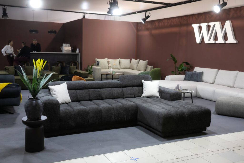 Duże stoisko z meblami tapicerowanymi. Na pierwszym planie ciemnoszara, pikowana sofa narożna z poduszkami. Z tyłu widać białe i beżowe sofy, a na ścianie duże, białe logo "WM".