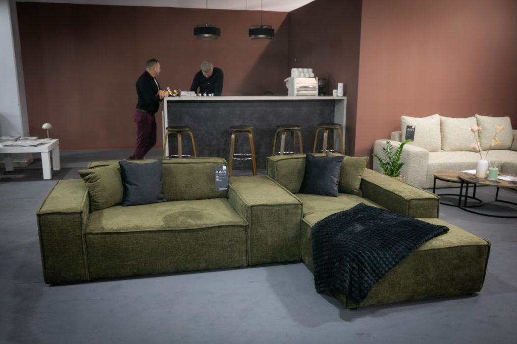 Modułowa sofa narożna w odcieniu oliwkowej zieleni z czarnym kocem i poduszkami, ustawiona na tle baru z wysokimi stołkami, przy którym stoją dwie osoby.