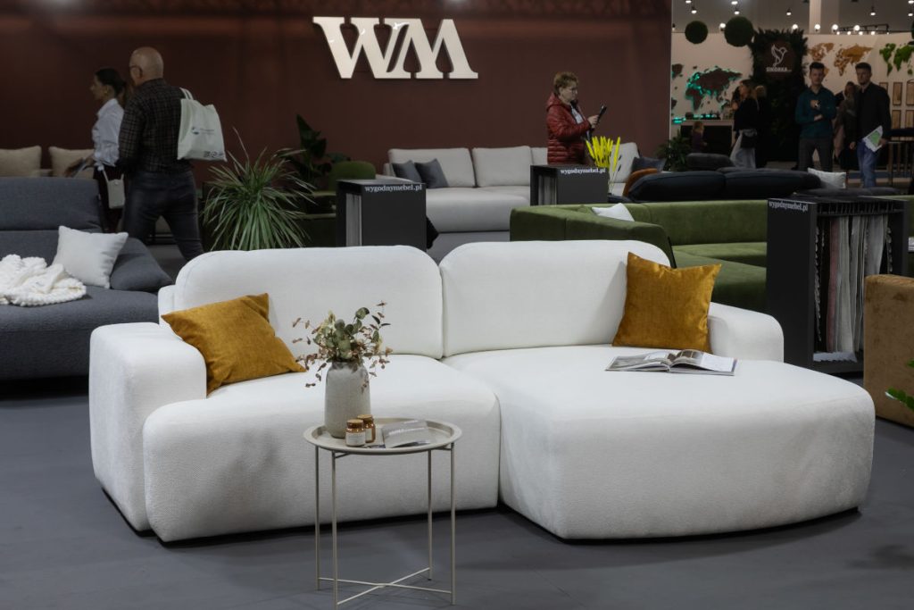 Detal białej, puszystej sofy modułowej z zaokrąglonymi kształtami i dwiema musztardowymi poduszkami. Obok mały, okrągły stolik z dekoracją. W tle widoczne jest duże logo "WM" na ścianie i inni zwiedzający.