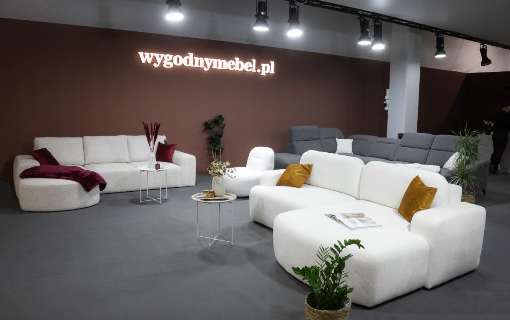 Przestronne stoisko targowe z nowoczesnymi, modułowymi sofami. Na pierwszym planie biała, puszysta sofa narożna w kształcie litery L z żółtymi poduszkami. Z tyłu widać drugą białą sofę oraz szare sofy w tle. Na ścianie logo "wygodnymebel.pl".