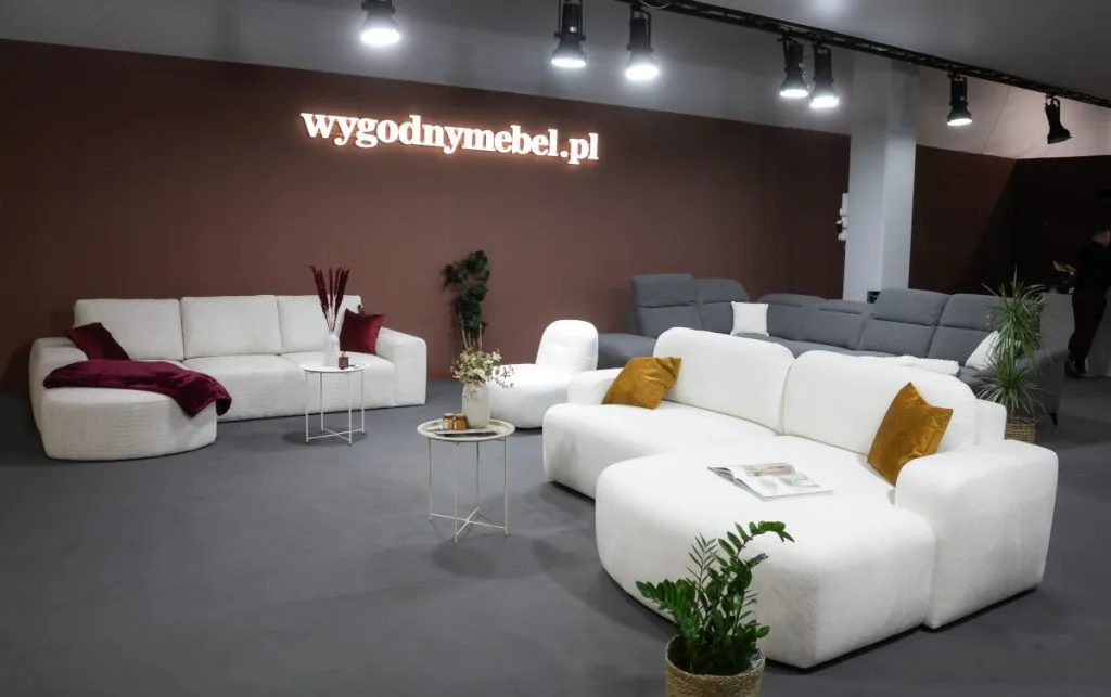 Przestronne stoisko targowe z nowoczesnymi, modułowymi sofami. Na pierwszym planie biała, puszysta sofa narożna w kształcie litery L z żółtymi poduszkami. Z tyłu widać drugą białą sofę oraz szare sofy w tle. Na ścianie logo "wygodnymebel.pl".
