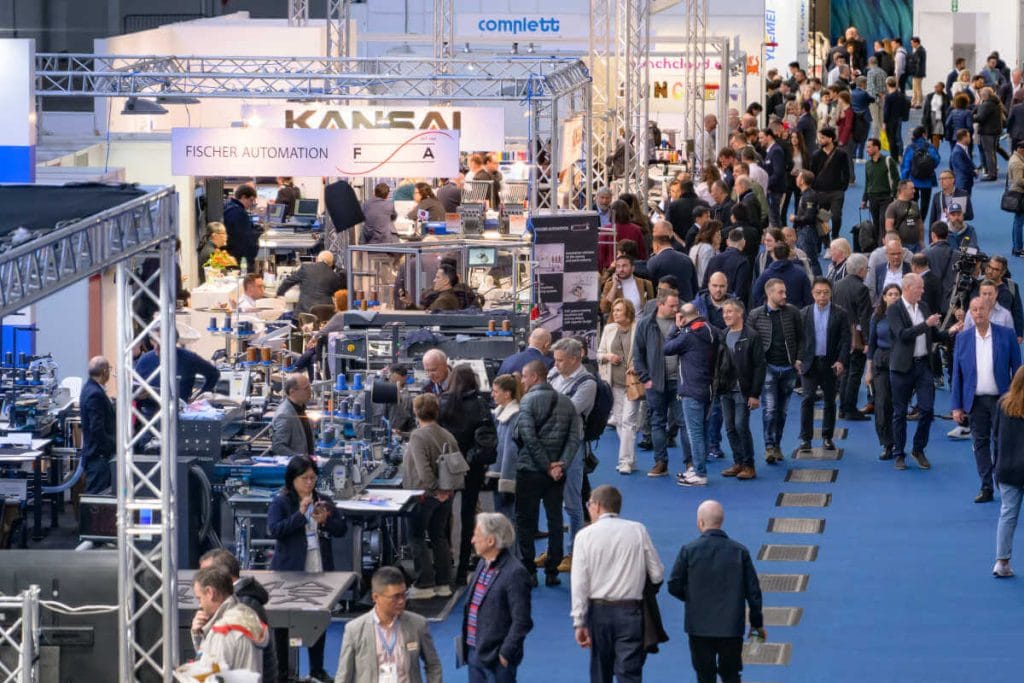 Szerokie ujęcie tłocznej hali targowej Texprocess z licznymi stoiskami wystawców, takich jak Kansai i Fischer Automation. Odwiedzający spacerują szeroką niebieską alejką, oglądając maszyny szwalnicze i urządzenia do obróbki tekstyliów.