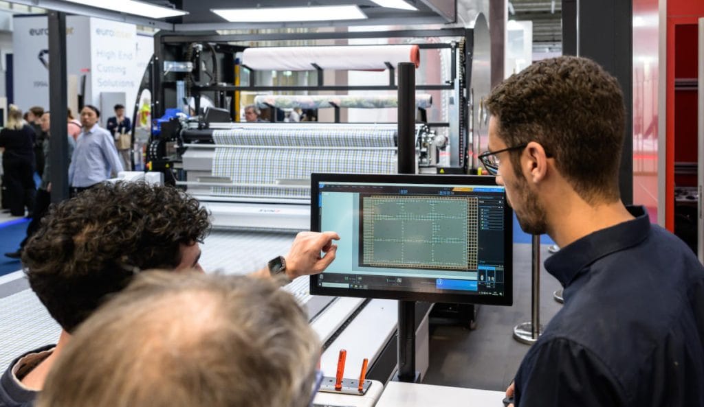 Trzech mężczyzn na targach branżowych analizuje projekt wykroju tkaniny wyświetlony na dużym monitorze dotykowym. W tle widoczna jest nowoczesna, zautomatyzowana maszyna krojcza z belką wzorzystego materiału.