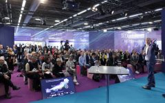 Prelegent w niebieskim garniturze przemawia na scenie podczas forum Techtextil 2024. Przed nim siedzi liczna publiczność na różowych pufach. Na podłodze widoczny jest ekran wyświetlający grafikę dotyczącą innowacji i zrównoważonego rozwoju.