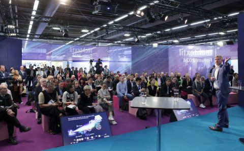Prelegent w niebieskim garniturze przemawia na scenie podczas forum Techtextil 2024. Przed nim siedzi liczna publiczność na różowych pufach. Na podłodze widoczny jest ekran wyświetlający grafikę dotyczącą innowacji i zrównoważonego rozwoju.