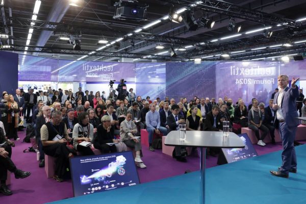 Prelegent w niebieskim garniturze przemawia na scenie podczas forum Techtextil 2024. Przed nim siedzi liczna publiczność na różowych pufach. Na podłodze widoczny jest ekran wyświetlający grafikę dotyczącą innowacji i zrównoważonego rozwoju.