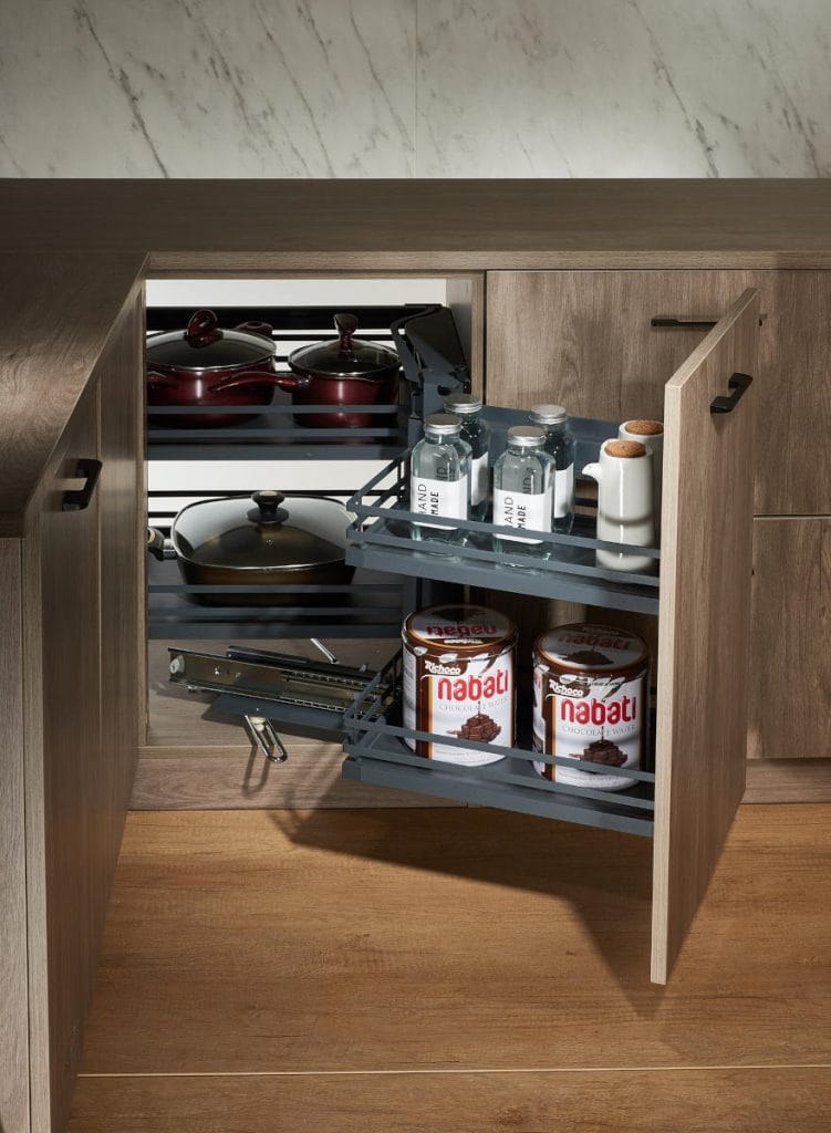 Zastosowanie systemu AMIX Corner Cargo w drewnianej szafce narożnej. Na wysuniętych, czarnych półkach znajdują się naczynia, garnki oraz produkty spożywcze.