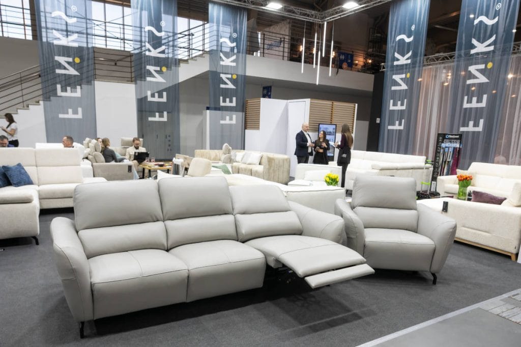 Ekspozycja jasnych mebli tapicerowanych na stoisku Feniks. Na pierwszym planie widać nowoczesną, szarą sofę z wysuniętym podnóżkiem (funkcja relaks) oraz pasujący do niej fotel. W tle widoczne są inne białe sofy, dekoracyjne tulipany oraz logotypy marki na pionowych banerach.
