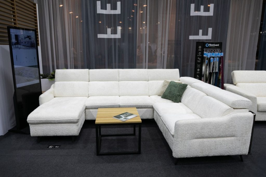 Duży narożnik o fakturze typu "bouclé" w kolorze złamanej bieli, udekorowany zielonymi poduszkami. Przed meblem stoi minimalistyczny stolik z drewnianym blatem na czarnym stelażu. Obok znajduje się stojak z wzornikami tkanin FibreGuard oraz pionowy ekran informacyjny.