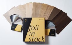 Żółto-czarny katalog Schattdecor „Foil in Stock” na tle rozłożonych próbek folii meblowych w różnych odcieniach drewna i szarości.