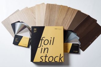 Żółto-czarny katalog Schattdecor „Foil in Stock” na tle rozłożonych próbek folii meblowych w różnych odcieniach drewna i szarości.