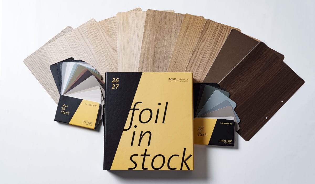 Żółto-czarny katalog Schattdecor „Foil in Stock” na tle rozłożonych próbek folii meblowych w różnych odcieniach drewna i szarości.