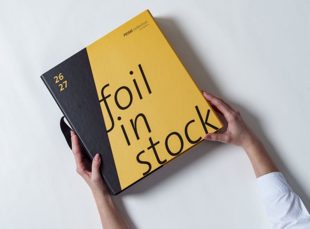 Zamknięty katalog Schattdecor z napisem „Foil in Stock” otoczony wachlarzem próbek oklein meblowych o strukturze drewna oraz gładkich dekorów.