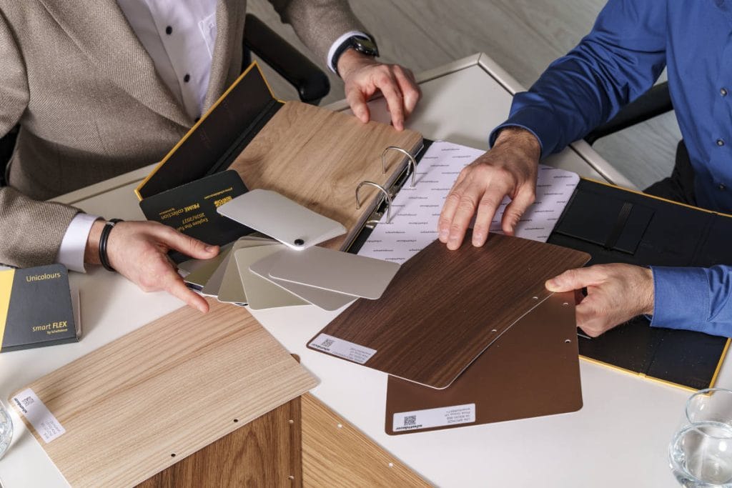 Dwie osoby przy białym stole analizujące wzornik folii Schattdecor; dłonie wskazują na próbki dekorów drewnopodobnych w jasnym i ciemnym odcieniu.