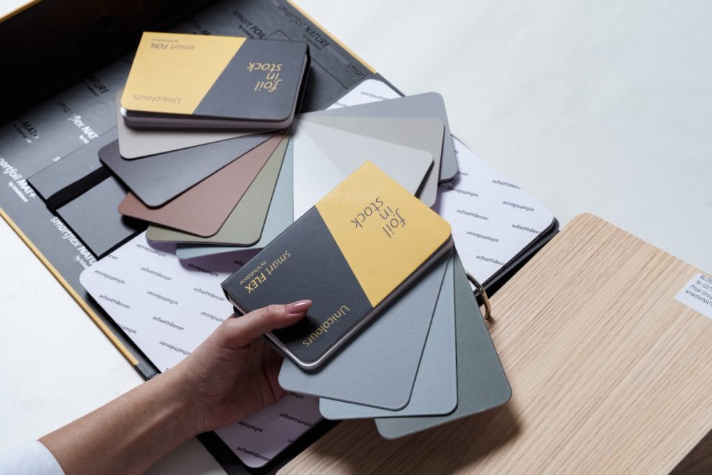 Zbliżenie na dłonie trzymające próbnik folii meblowych Schattdecor z kolekcji Foil in Stock; widoczne różne warianty kolorystyczne od szarości po dekory drewniane.