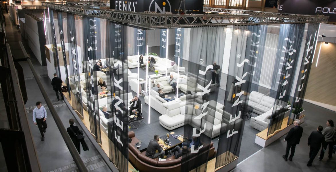 Widok z góry na przestronne stoisko wystawowe marki Feniks na targach Meble Polska 2026. Stoisko jest ogrodzone półprzezroczystymi, ciemnymi zasłonami z białym logo firmy. Wewnątrz znajdują się liczne zestawy wypoczynkowe, sofy i narożniki, przy których siedzą i rozmawiają ludzie.