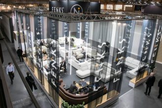 Widok z góry na przestronne stoisko wystawowe marki Feniks na targach Meble Polska 2026. Stoisko jest ogrodzone półprzezroczystymi, ciemnymi zasłonami z białym logo firmy. Wewnątrz znajdują się liczne zestawy wypoczynkowe, sofy i narożniki, przy których siedzą i rozmawiają ludzie.