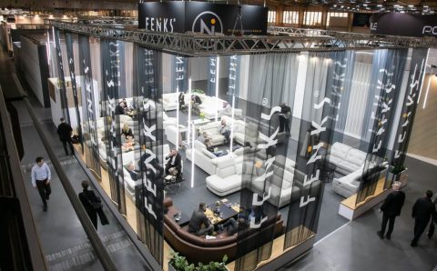 Widok z góry na przestronne stoisko wystawowe marki Feniks na targach Meble Polska 2026. Stoisko jest ogrodzone półprzezroczystymi, ciemnymi zasłonami z białym logo firmy. Wewnątrz znajdują się liczne zestawy wypoczynkowe, sofy i narożniki, przy których siedzą i rozmawiają ludzie.