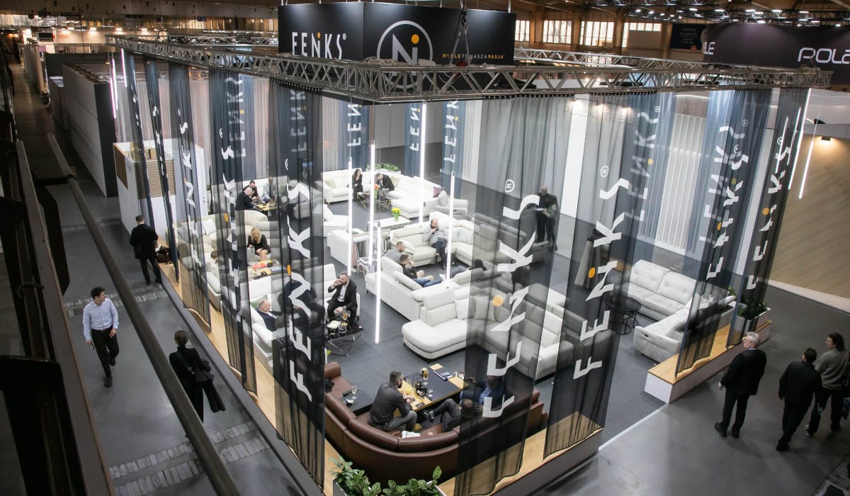 Widok z góry na przestronne stoisko wystawowe marki Feniks na targach Meble Polska 2026. Stoisko jest ogrodzone półprzezroczystymi, ciemnymi zasłonami z białym logo firmy. Wewnątrz znajdują się liczne zestawy wypoczynkowe, sofy i narożniki, przy których siedzą i rozmawiają ludzie.