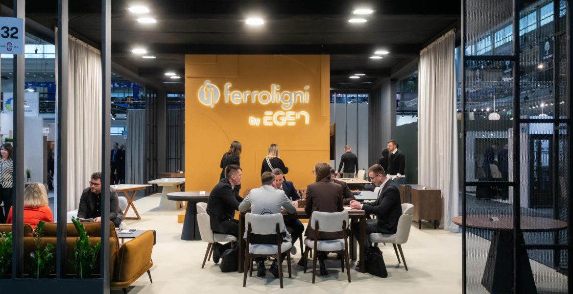 Widok na wnętrze stoiska Ferroligni by EGEN na targach Meble Polska. W centralnym punkcie znajduje się grupa osób rozmawiających przy stole, w tle duża pomarańczowa ścianka z podświetlanym logo firmy.