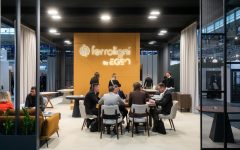 Widok na wnętrze stoiska Ferroligni by EGEN na targach Meble Polska. W centralnym punkcie znajduje się grupa osób rozmawiających przy stole, w tle duża pomarańczowa ścianka z podświetlanym logo firmy.