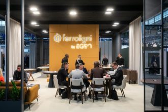 Widok na wnętrze stoiska Ferroligni by EGEN na targach Meble Polska. W centralnym punkcie znajduje się grupa osób rozmawiających przy stole, w tle duża pomarańczowa ścianka z podświetlanym logo firmy.